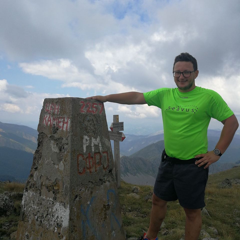 Igor smiling beside a mountain summit marker, with mountains and clouds behind him.